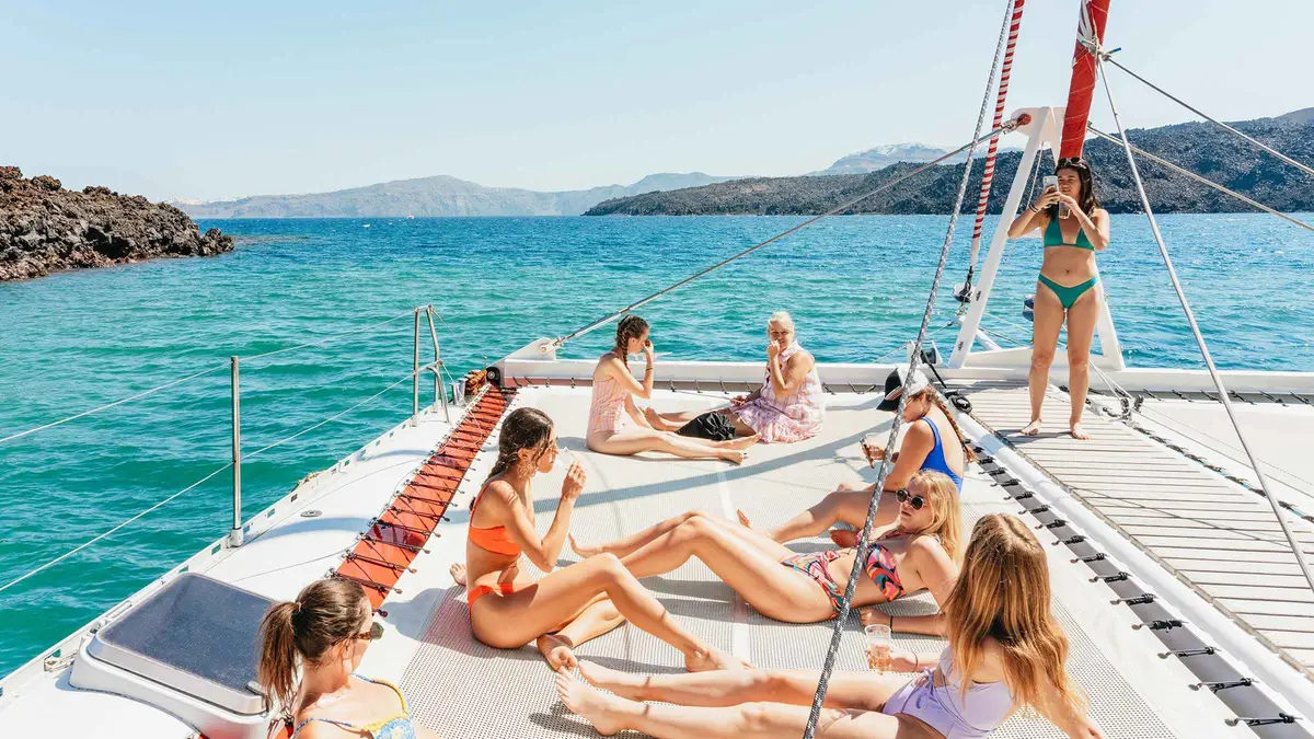 White catamaran with guests at the rail sailing past the red volcanic cliffs during a Santorini boat tour in the caldera, Greece